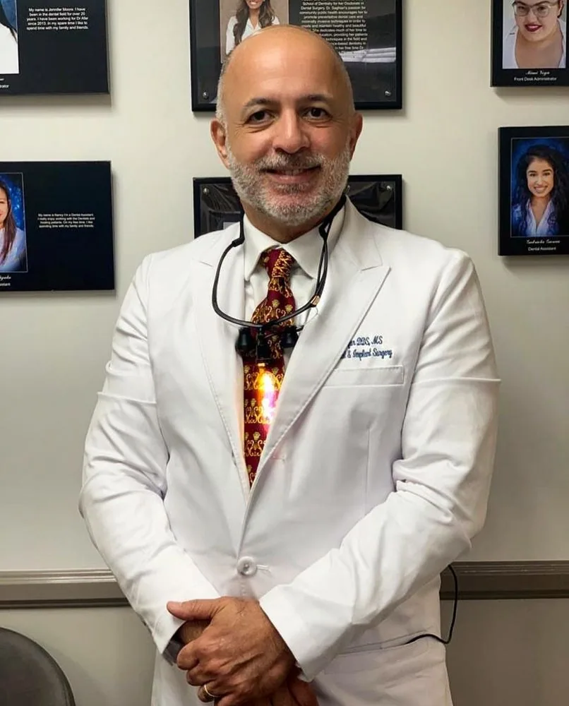 Dr. Bijan Afar standing in a white coat in front of the office team photo wall