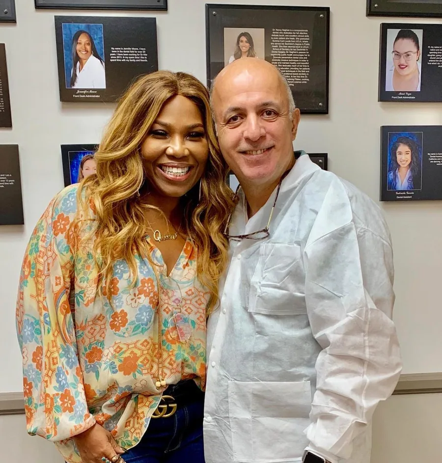 Dr. Bijan Afar smiling next to a patient in front of the office team photo wall
